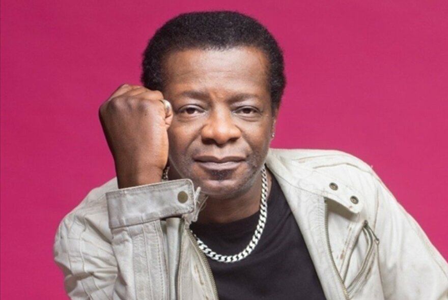 Stephen K Amos sitting with his fist next to his head, wearing a light coloured jacket and silver necklace.