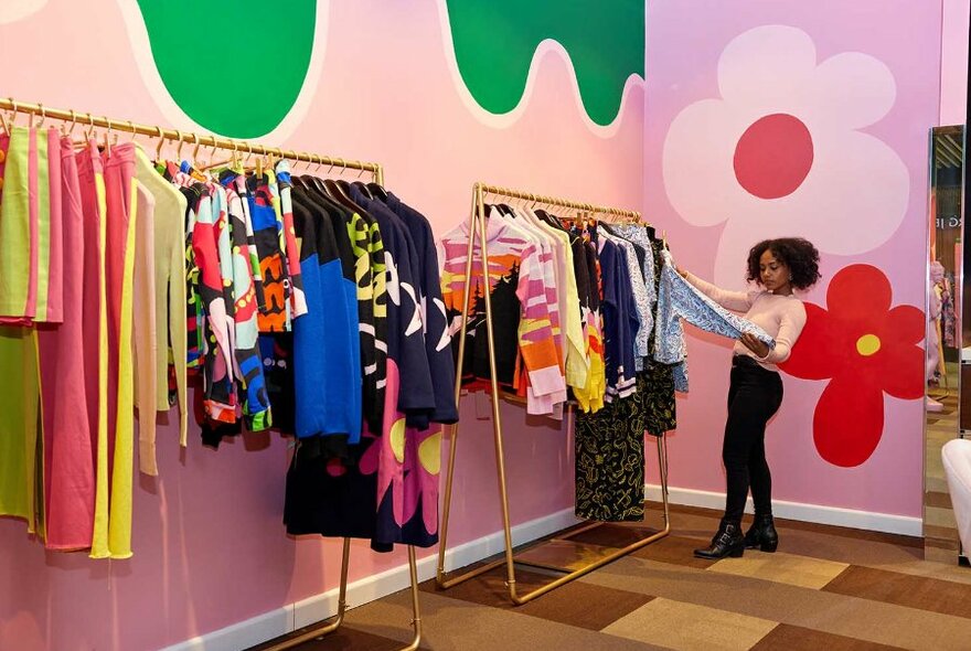 A woman browsing in a store with colourful clothing and a floral mural on the wall. 