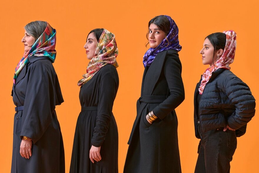 Four performers in a row, in profile, all wearing black garments and head scarfs, standing against an orange background.