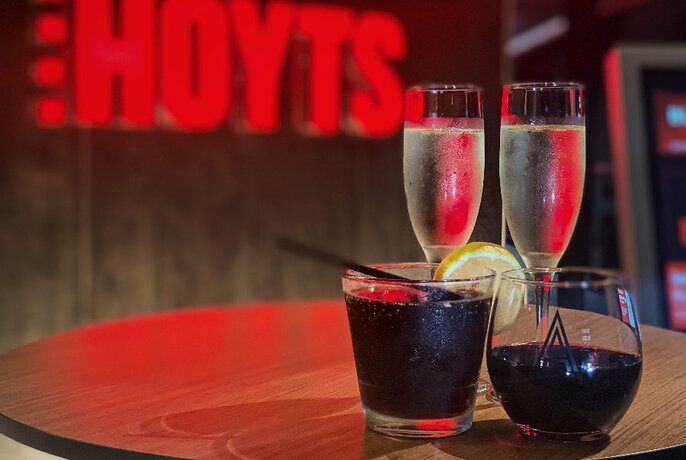 Two glasses of sparkling wine and a couple of other drinks on a table with a HOYTS neon sign in the background.