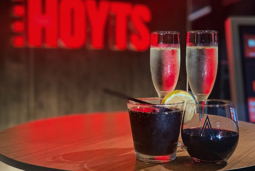 Two glasses of sparkling wine and a couple of other drinks on a table with a HOYTS neon sign in the background.
