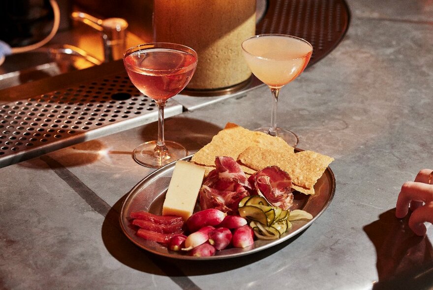 A plate of antipasti nibbles including pickled vegetables, cured meats and cheese on a tabletop with two small cocktails next to it.