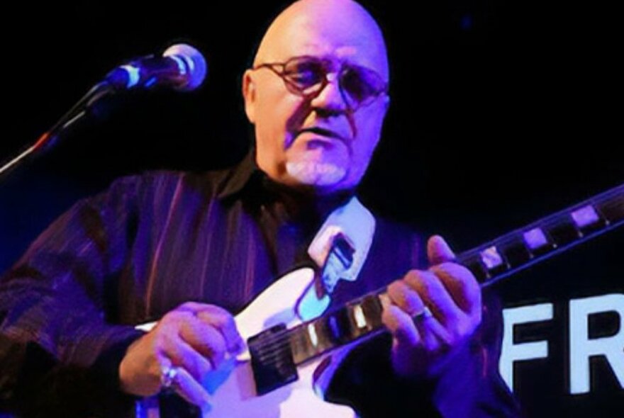 Guitarist Frank Gambale playing on stage in front of a microphone with a purple stage light on him. 