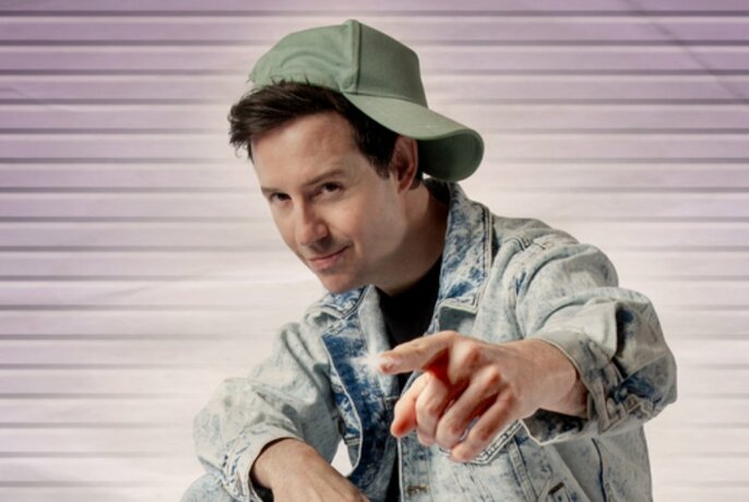 Comedian Nath Valvo looking comical in cap turned sideways, acid wash denim jacket and a finger pointing straight ahead.