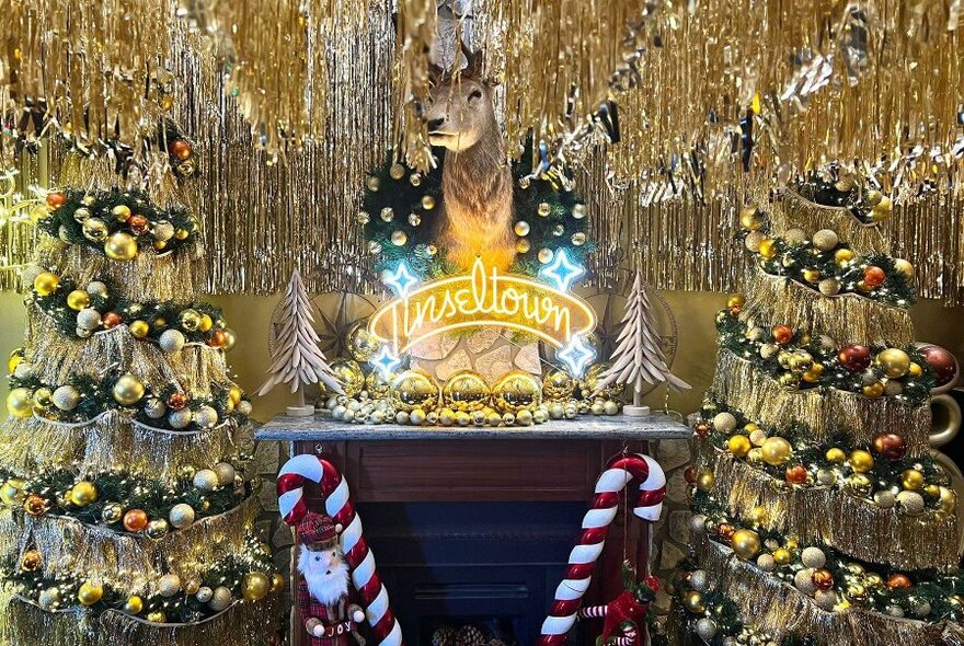 A Christmas themed wall of gold decorations, including a wall mounted reindeer, Christmas trees with baubles, tinsel, and oversized red and white striped candy canes.