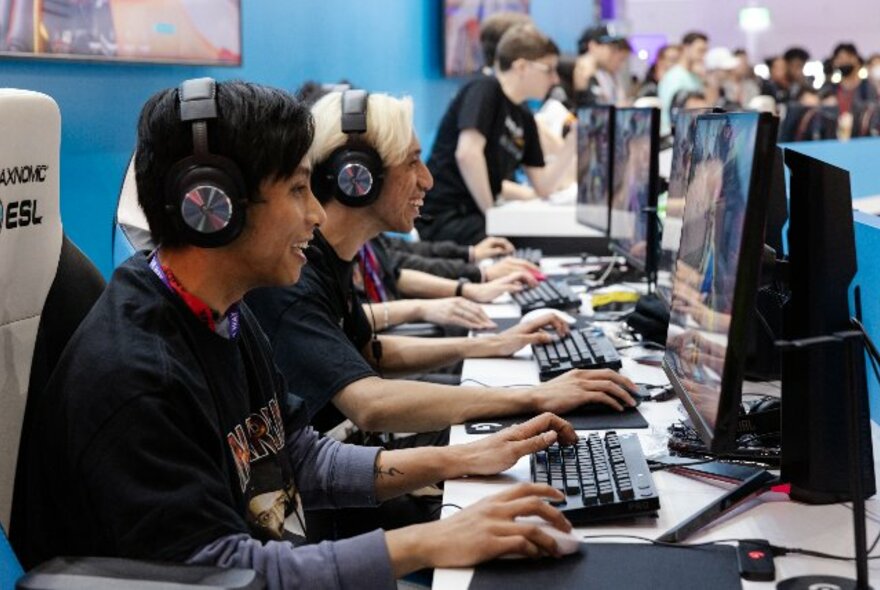 People wearing headphones seated at computers on a long table in a crowded exhibition hall.