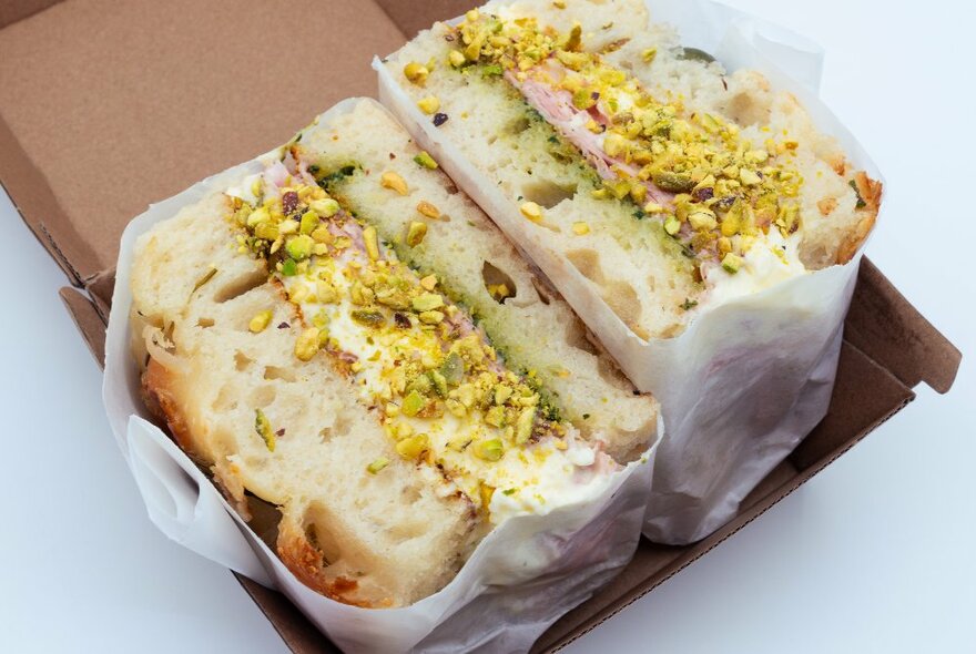 Wrapped focaccia sandwich in a cardboard box with creamy white filling and chopped pistachios.