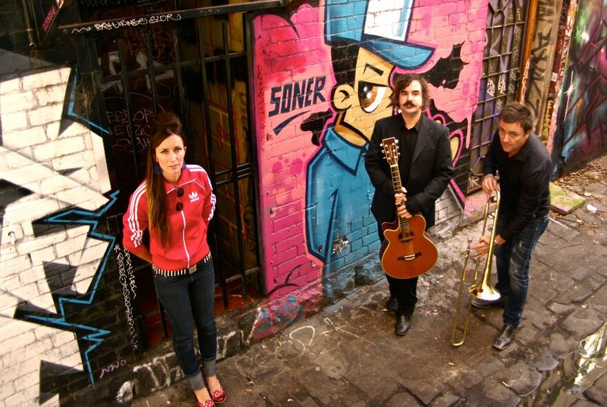 Two men carrying musical instruments, and a woman, stand in an outdoor laneway in front of a wall covered in street art.