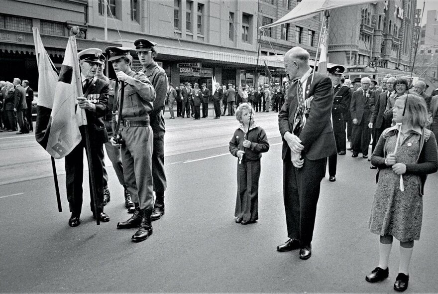 An Anzac Day street parade in the centre of Melbourne city, with veterans, soldiers in uniform and young children walking side-by-side, some holding flags. 