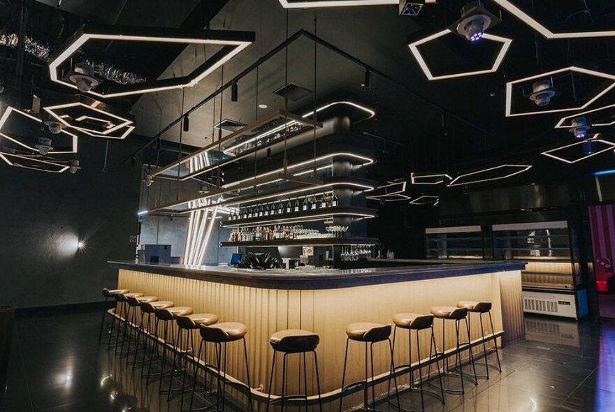 Interior of a bar, with dark ceilings, walls and floor, a central bar area with high bar stools under the counter.