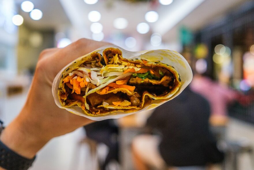 A person is holding a savoury Chinese crepe filled with vegetables and meat