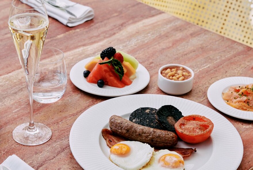 A cooked breakfast plate, plate of fruit and a glass of bubbles on a table top.