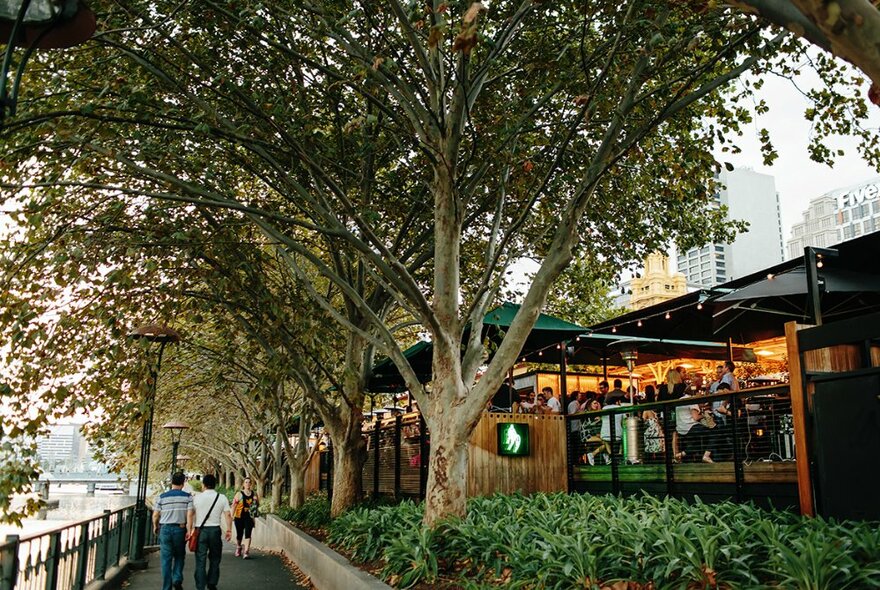 Exterior of Arbory Bar building, showing the wooden outdoor deck, situated on the banks of the Yarra River, surrounded by green leafy trees.