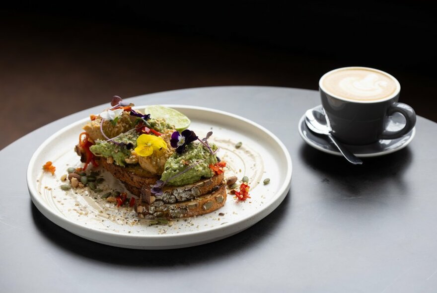 A brunch dish of smashed avocado on toast with edible flower garnish and a flat white coffee.