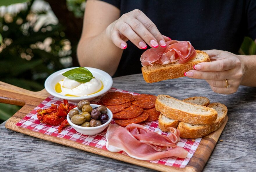 A hand taking prosciutto from an antipasto platter and placing it on a slice of toasted bread.