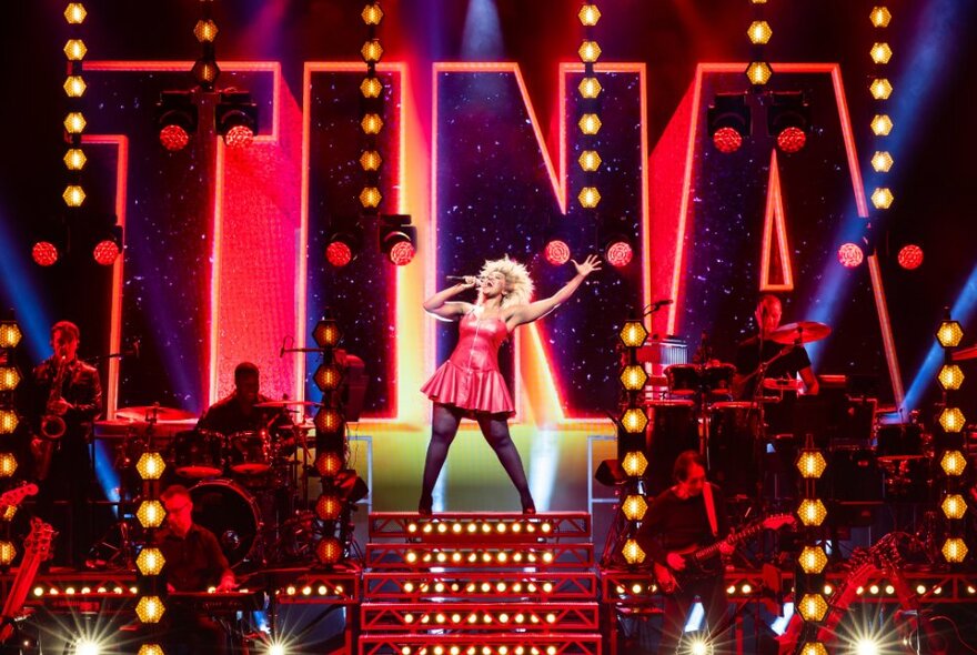 Performer dressed as Tina Turner, singing on a podium with huge neon lights behind her.