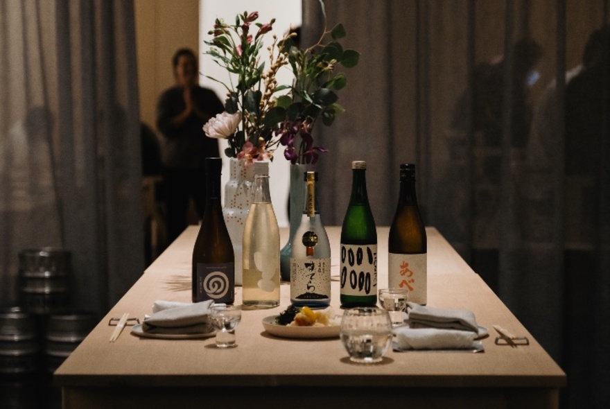 Table set for a dinner service with two plates, napkins, cutlery, a floral arrangement and five different bottles of sake.