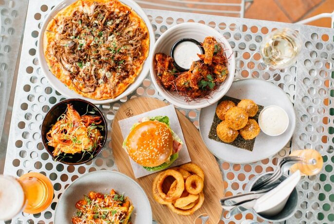 Overhead view of a table with plates of food including a pizza, burger and onion rings, and plates of pasta.