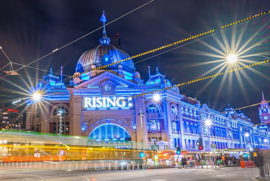 Flinders train station at a busy intersection lit up in blue with the words 'RISING' on the front