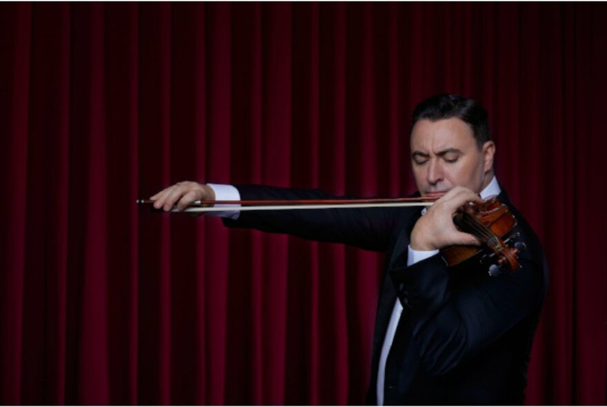 Man in a tuxedo playing a violin, standing in front of a red velvet curtain. 