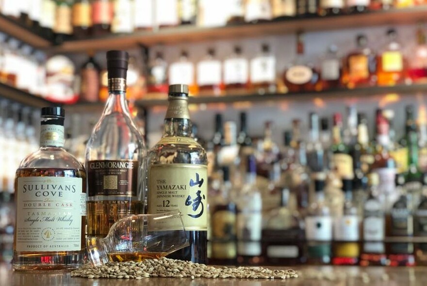 Bar counter with three bottles of whisky and a glass on its side on a handful of grain, with blurred bottles in the background.