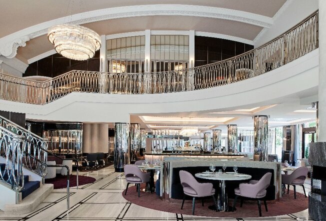 A hotel lounge with a sweeping staircase, chandelier and mirrored poles.