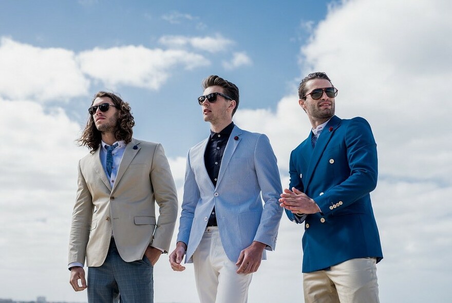 Three male models outside against blue sky with clouds, wearing smart-casual jackets and sunglasses.