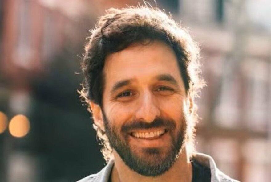 Smiling head shot of a man with a brown curly short hair, and a beard, standing outdoors with blurred buildings behind him.