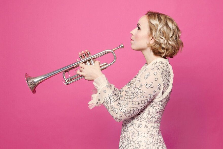 A woman in a white patterned dress stands in profile holding a trumpet almost to her mouth against a hot pink background. 