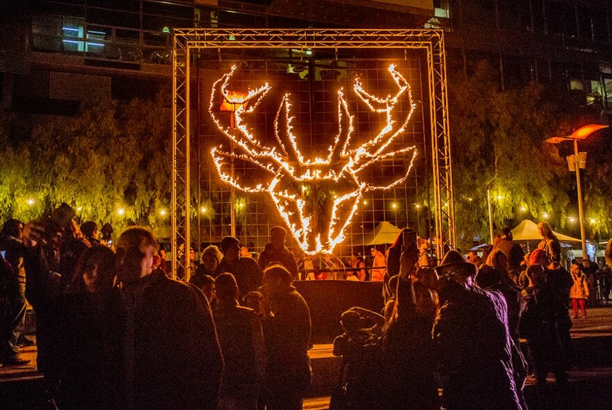 Crowds gathered around a large flaming deer head artwork at an outdoor festival.