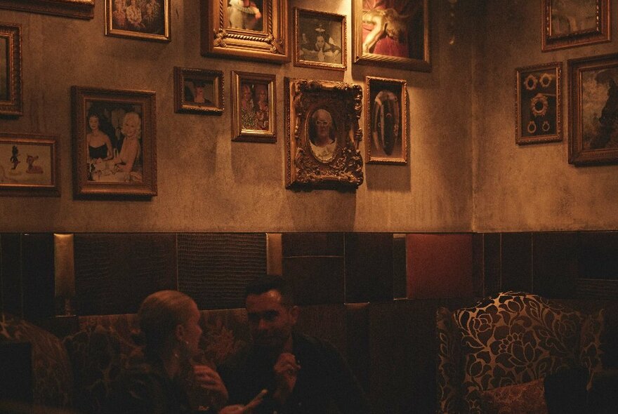 Dimly lit interior of Gin Palace, small selection of framed paintings on a wall, and two people sitting on a couch and sipping drinks.