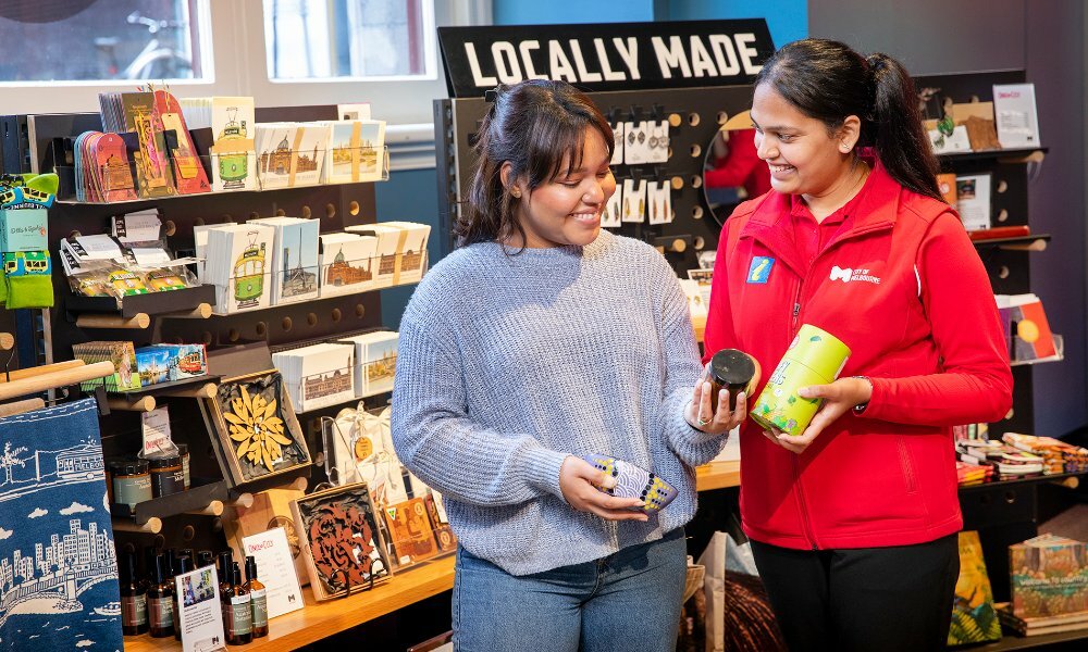 A volunteer in a tourist info centre showing gift products to a smiling customer.