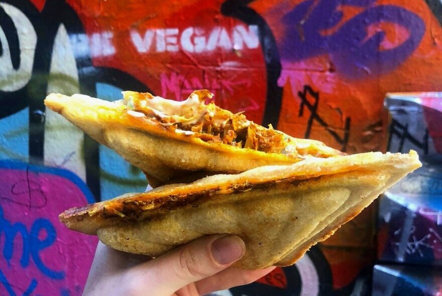 A hand holding a cheesy vegan jaffle in front of a colourful graffiti wall.