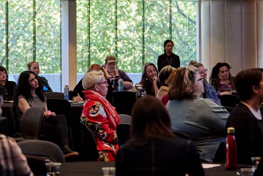 People at a symposium looking at a speaker in a room with long windows looking out to greenery. 