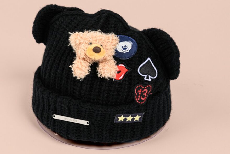 A black beanie with ears, patches and a small teddy bear attached.