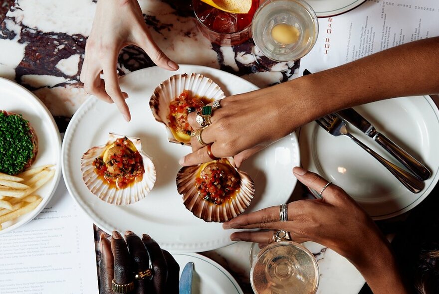Two sets of hands reaching in to select food from a plate on a restaurant table, drinks and cutlery also on the table.
