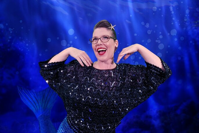 Ethel Mermaid smiling and flapping her arms, wearing a star in her hair and standing in front of a blue background.
