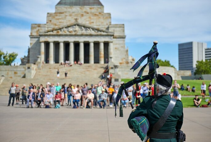 People gathered in front of the Shrine of Remembrance and a man playing bagpipes with his back to the camera.