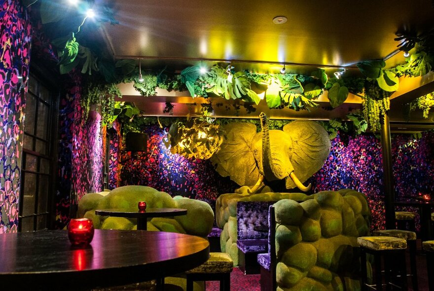 Interior of a club with ornate jungle themed decor featuring large green plants, an elephant sculpture hanging from the wall and booth seating.