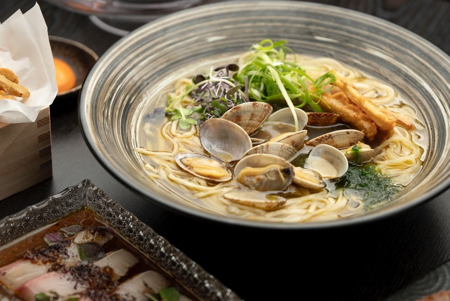 A bowl of Japanese clam ramen in a traditional japanese bowl, with a plate of sashimi in the foreground.