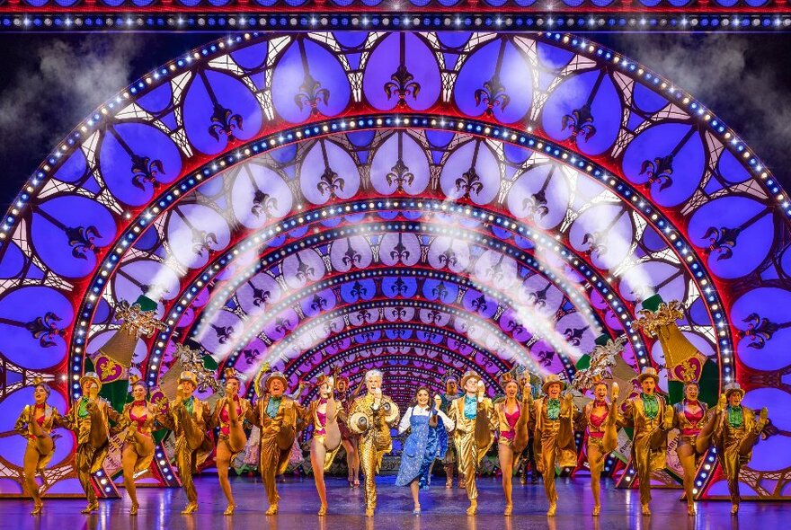 The cast of Beauty and the Beast in their costumes on a large theatre stage, kicking up their legs in a chorus style line-up, against a blue and purple stained glass style backdrop.