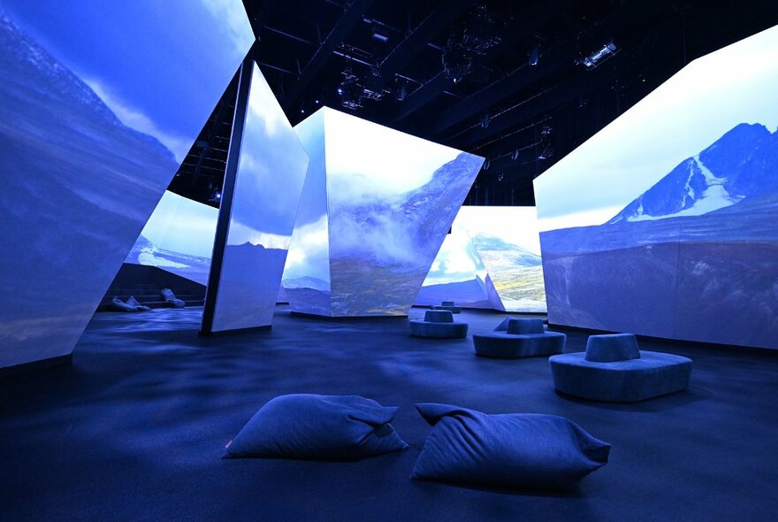 Large digital panels displaying artic style landscapes in a darkened gallery space with benches and beanbags on the ground below for people to sit on while viewing.