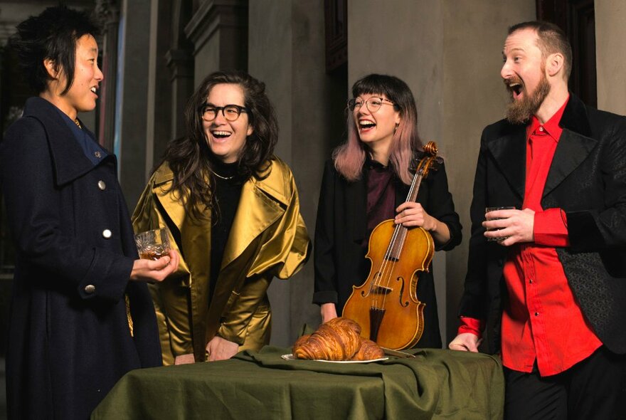 Musical quartet smiling and laughing, one holding a violin, in front of a plate of croissants.