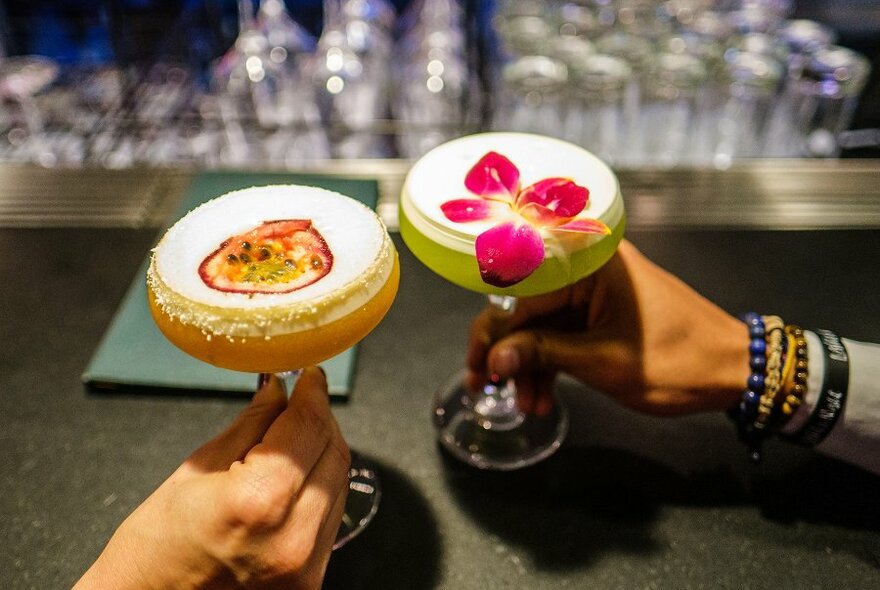 Two people holding colourful foamy cocktails with a flower and passionfruit garnish.