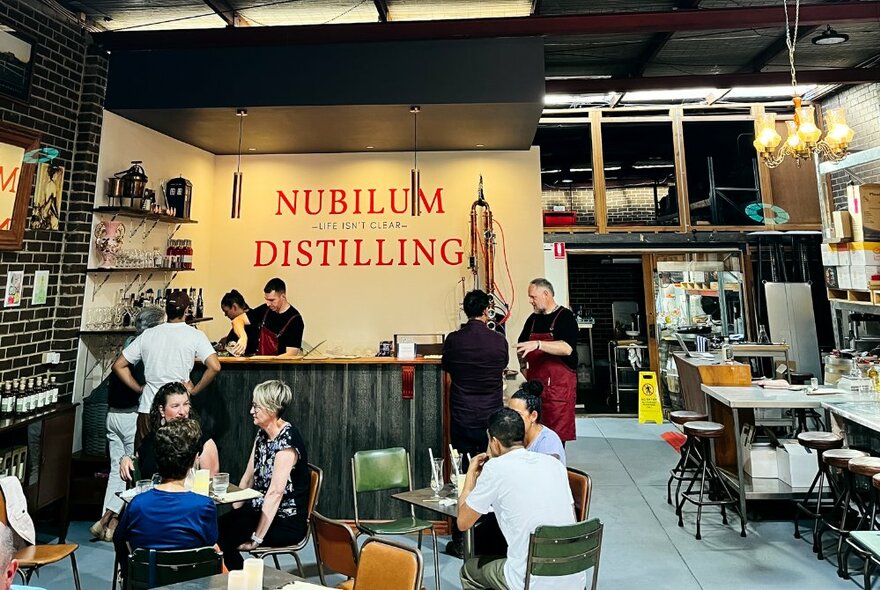 The interior shed space of Nubilum showing patrons sitting at tables and chairs socializing and drinking, a small bar area and distilling equipment  in the rear.