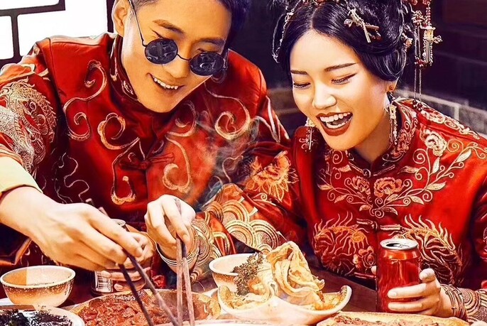Couple in traditional Chinese costumes smiling and selecting food from a plate with chopsticks.