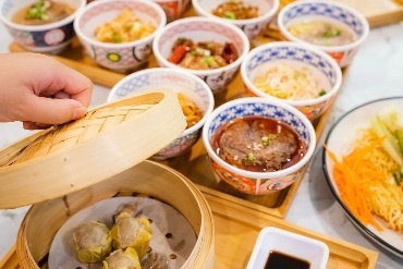 A hand lifting the lid on a serving of dumplings, next to a tray loaded with 12 dishes of food in Chinese bowls.