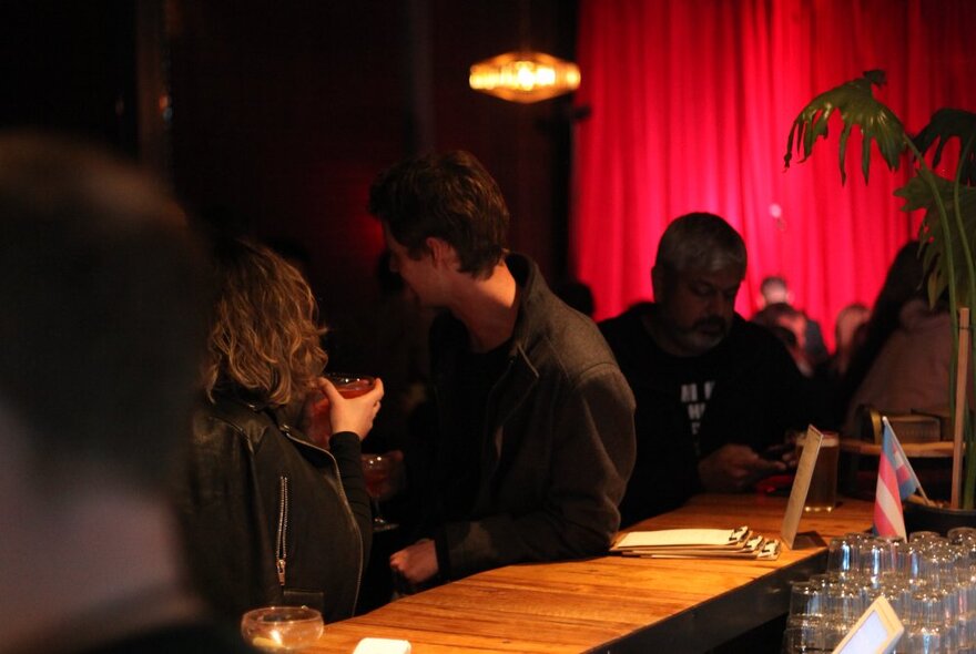 Two people leaning against a wooden bar, sipping drinks, soft lights and a red curtain in the background.