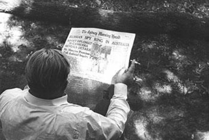 Rear view of a person reading the front of a broadsheet newspaper, with a cigarette in their hand, outdoors, black and white images from the mid-1950s.