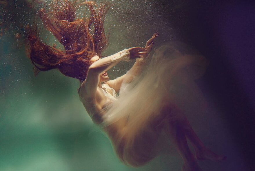 A red headed woman with long hair in a gauzy dress sinking backwards under the water highlighted by rays of light.